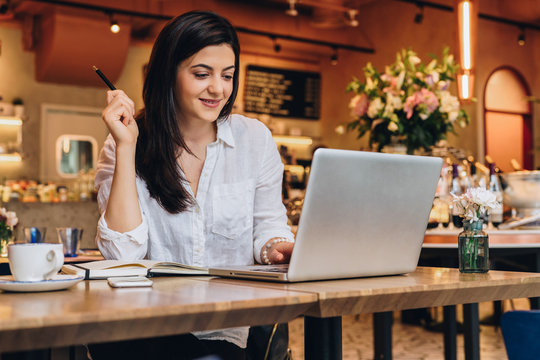 Young businesswoman wearing in shirt sitting in cafe at table, looking on screen of computer and smiling. Freelance, startup. Distance work, learning, blogging. Online marketing, education. Lifestyle.
