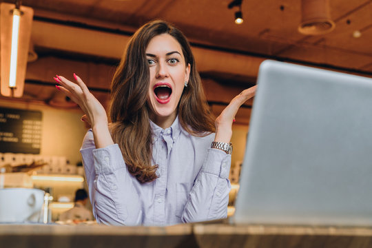 Young businesswoman is sitting in cafe at table in front of laptop and opening her mouth and raising her hands in surprise looks at screen. Girl received an email and is happy with good news.