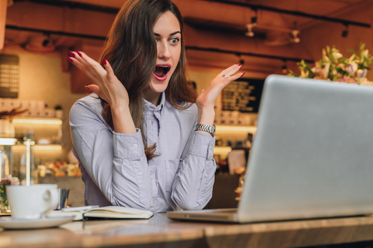 Young businesswoman is sitting in cafe at table in front of laptop and opening her mouth and raising her hands in surprise looks at screen. Girl received an email and is happy with good news.