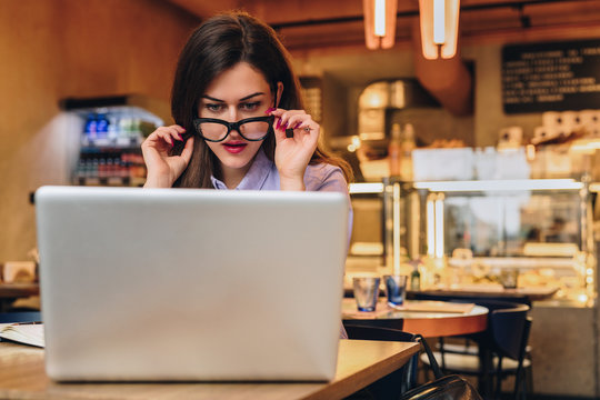 Front view. Young business woman is sitting in cafe at table in front of laptop and looks at screen in surprise, lowering her glasses. Hipster girl blogging, learning online. Distance work, education.