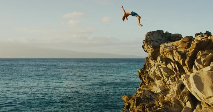 Young man doing backflip cliff jumping into ocean at sunset