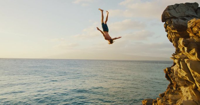 Young man doing backflip cliff jumping into ocean at sunset