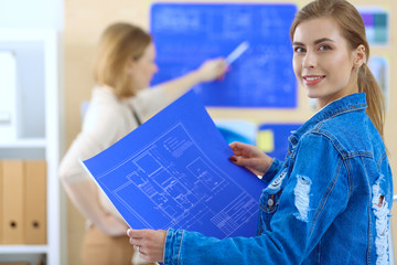 Two young woman standing near desk with instruments, plan and laptop.