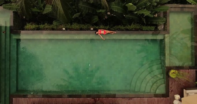 Female in bikini relaxing on the edge of the swimming pool and enjoying her vacation. Aerial view of young woman lying on the poolside of luxury resort.
