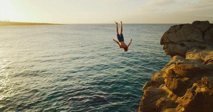 Young man doing backflip cliff jumping into ocean at sunset