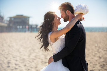 Beautiful bride and handsome groom embrace and kiss at beach during sunset along ocean pier