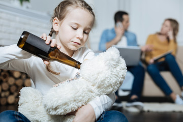 Beverage. Unhappy fair-haired little girl holding a bottle of beer and giving it to her teddy bear while her parents working on their laptops
