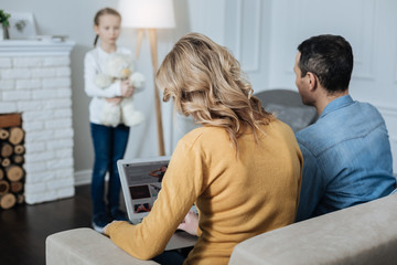 Obraz premium Play with me. Lonely slim little girl holding her teddy bear and standing in front of her parents while they working on their laptops