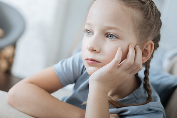 Thoughtful. Concentrated fair-haired blue-eyed little girl thinking and looking in the distance while sitting on the sofa