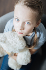 Feeling alone. Unhappy blue-eyed little girl hugging and holding her teddy bear while sitting in the armchair