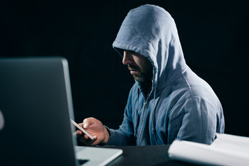 male hacker in a sweatshirt with a hood sits at the table and looks into the phone