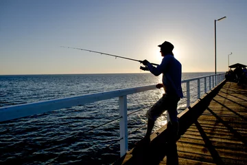 Fototapeten silhouette of fisherman with hat and fish rod standing on sea dock fishing at sunset with beautiful orange sky in vacations relax hobby © Wordley Calvo Stock