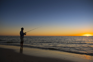 silhouette of fisherman with hat on the beach with fish rod standing on sea water fishing at sunset with beautiful orange sky in vacations
