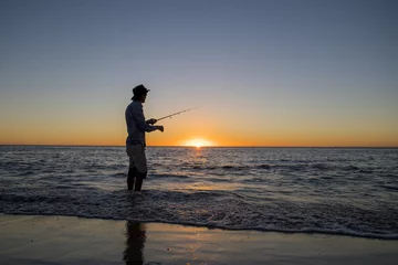 Fotobehang silhouette of fisherman with hat on the beach with fish rod standing on sea water fishing at sunset with beautiful orange sky in vacations © Wordley Calvo Stock