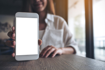 Mockup image of a woman holding and showing white mobile phone with blank screen on the table in modern cafe