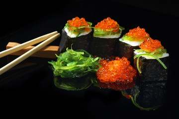 Sushi with caviar and algae salad next to chopsticks on a black background with reflection.