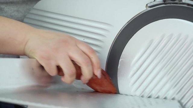 Electric food slicer. Meat slicing machine cutting processed meat products. Stainless steel blade slices smoked and cured pork bacon. Woman chef cuts pork loin.