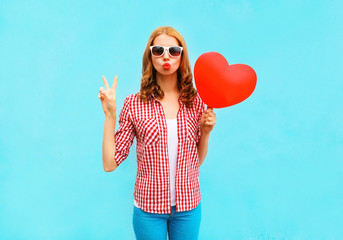 Pretty woman makes an air kiss with red balloon in the shape of a heart on blue background