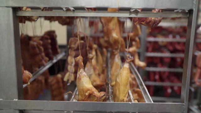Meat and sausage manufacturing. Smoked chicken and pork bacon hanging on racks in storage room at meat processing factory. Processed meat products in refrigerated warehouse of sausage making plant.