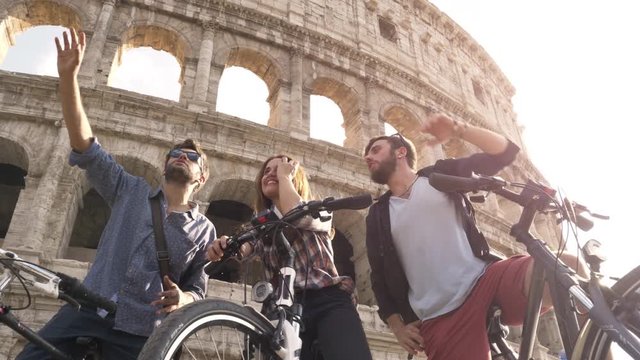 Three happy young friends tourists with bikes and backpacks at Colosseum in Rome having fun talking taking pictures on sunny day slow motion ground shot