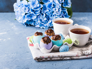 Easter sweet cup cakes treets in colorful pastel egg shells with chocolate and hydrangea flowers on blue background. Festive holiday home made food with cups of tea