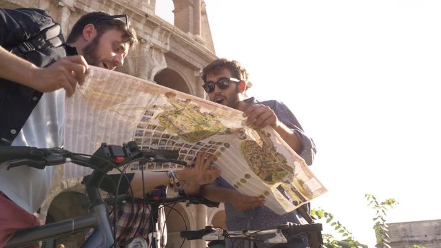 Three happy young friends tourists with bikes and backpacks at Colosseum in Rome reading map guide for directions on sunny day slow motion steadycam ground shot