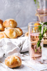 Easter table setting with colored orange eggs, plate of hot cross buns, green branches decorated, glass of cocktail drink over white plank wooden table with textile tablecloth.