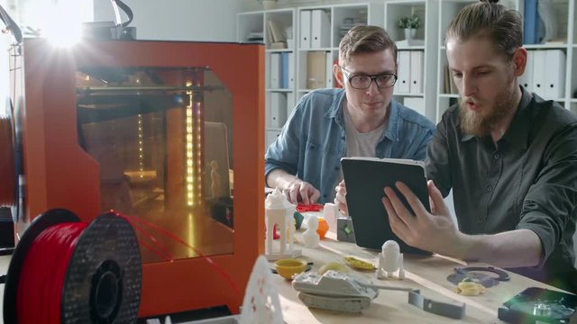 Two young men controlling 3D printing process via digital tablet while creating 3D print models in designer studio