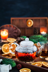 Glass teapot of hot black tea on cozy background with dried oranges and candles
