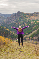 Naklejka na ściany i meble A girl in a lilac jacket stretching her arms on a mountain, a view of the mountains and an autumn forest by a cloudy day, free space for text
