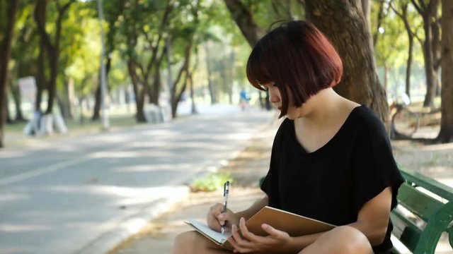 Happy hipster young asian woman writing into her diary in park. Happy hipster young asian woman working on notebook in park. Student studying outdoors. Lifestyle woman outdoor concept.