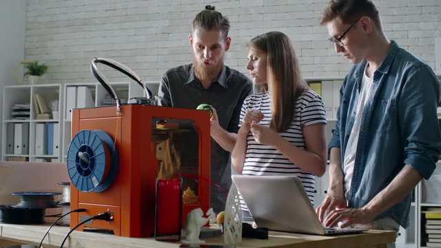 Tracking shot of young man and woman talking about 3D printing process while their male colleague controlling 3D printer via laptop