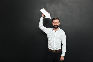 Good-looking brunette man holding blank speech arrow pointer directing towards him, over dark gray wall copy space