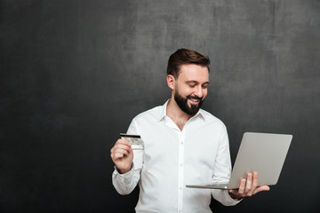 Portrait of cheerful man doing online payment in internet using notebook and credit card, isolated over dark gray background