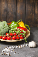 Diet, Healty Food Cooking, Vegetarian Concept. A plate of fresh raw vegetables: cherry tomatoes, broccoli, paprika peppers, rosemary, garlic and spices on a dark table against a wooden wall