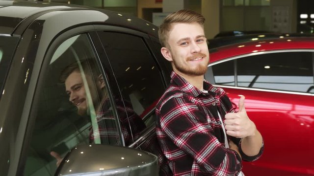 Handsome man showing thumbs up leaning on a new car at the dealership