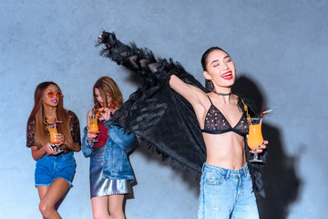 cheerful stylish asian girl holding glass with cocktail while friends drinking behind at party