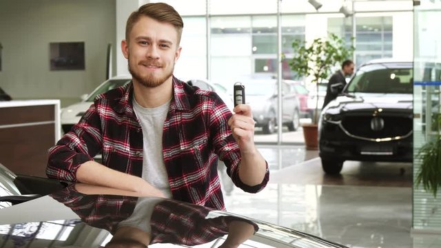 Young handsome man showing car keys to his new auto at the dealership salon
