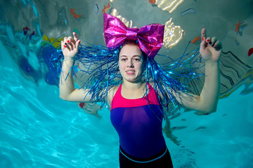 Fototapeta na wymiar Fun girl swims underwater in the pool, arms outstretched to the sides, with a big red bow on her head, looks at camera and smiling. Portrait. Horizontal orientation. The view from the bottom
