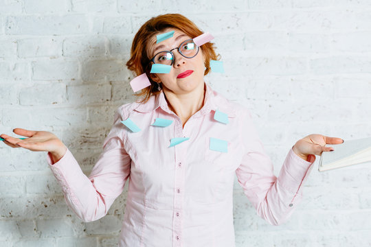 Red hair comic woman in glasses with stickers on her, looking at camera with a quizzical lose courage emotion, and writting in notebook a lot of things to do. White brick wall with shadow behind her.