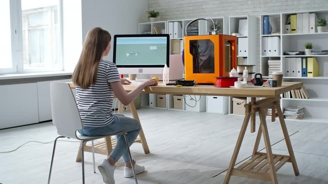 Rear view of young female designer sitting at desk in the office and creating 3D digital design on computer for 3D printer