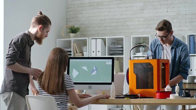 Team of young engineers working together in studio: woman drafting 3D model on computer and speaking with male colleague while man watching how 3D printer works