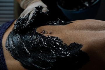 Young woman getting spa treatment in salon. Beautiful girl having clay body mask apply on belly by beautician. Mud therapy. Close up
