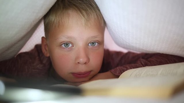 Closeup portrait of smiling laughing little 10 years old kid looking at camera. Boy reads paper book laying in his bed at night. Kid hiding under white blanket and uses lamp of telephone for lighting.