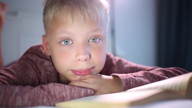 Closeup portrait of cute little kid in his bed rubbing his tired eye with hands after long reading of interesting paper book.