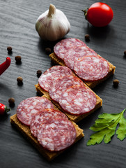 Tasty appetizer sandwich of thinly sliced spicy salami on bread on a dark wooden background