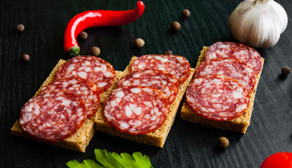 Tasty appetizer sandwich of thinly sliced spicy salami on bread on a dark wooden background