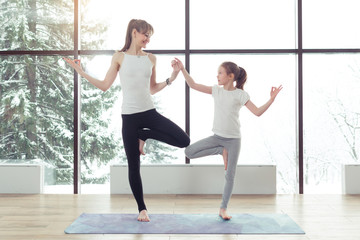 Obraz premium Young beautiful woman and charming little daughter are doing difficult yoga exercises and smiling while working out at gym with beautiful scenery outside window with sunlight. Healthy fitness concept
