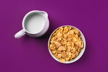 Bowl with corn flakes, jug of milk on purple background, top view