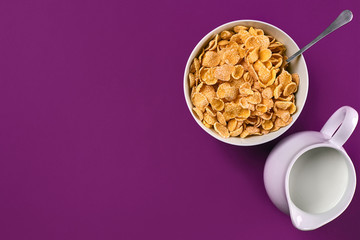 Bowl with corn flakes, jug of milk and spoon on purple background, top view